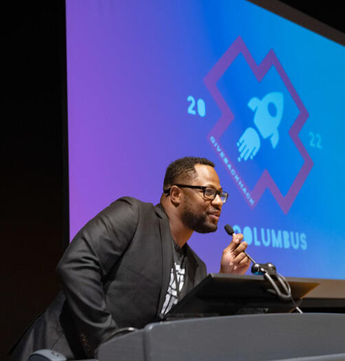 A photo of GiveBackHack organizer Clayton Nelson at the podium in front of an event slide design.